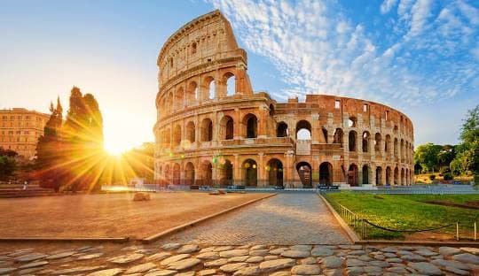The Colosseum in Rome at sunrise with golden sunlight illuminating the ancient amphitheater and cobblestone plaza